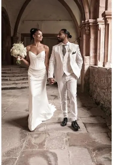 Die Braut und der Bräutigam spazieren Hand in Hand durch die Gänge eines historischen Gebäudes, sie tragen ein stilvolles Hochzeitskleid und einen eleganten Anzug.